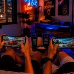 A imagem mostra duas pessoas sentadas em um sofá jogando um jogo multiplayer de carros rebaixados em seus smartphones. Cada celular exibe um carro personalizado em um mapa urbano inspirado no Brasil, enquanto o ambiente gamer conta com iluminação RGB, decoração moderna e uma vista noturna, transmitindo uma atmosfera descontraída e imersiva para partidas online entre amigos
